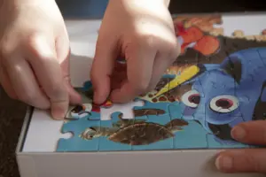 Kind setzt mit einer erwachsenen Person ein buntes Kinderpuzzle auf dem Teppich zusammen